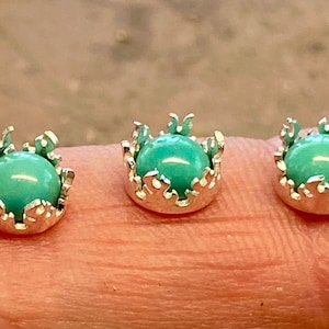 May include: Five small stud earrings, each featuring a smooth, round, light turquoise-coloured stone set within a silver-toned metal crown-shaped bezel. The earrings are arranged in a horizontal row, showcasing the detailed prong settings.