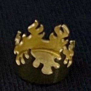 May include: A gold-coloured crown-shaped pin with a dark green base. The pin features a stylised design with multiple points and a curved base. The crown is set against a dark blue background.