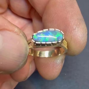 May include: A gold ring featuring an oval-shaped opal gemstone with vibrant blue and green flashes. The opal is set in a silver-toned bezel with notched edges, mounted on a textured gold band that splits into two sections near the setting.