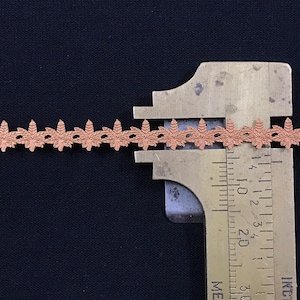 May include: A close-up of a brown lace trim measured with a ruler. The trim is approximately 1.2 cm wide and features a delicate floral design.