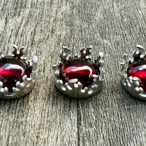 May include: Five silver-coloured, crown-shaped pendants, each featuring a deep red, round gemstone. They are arranged in a row on a textured, grey wooden surface. The gemstones have a polished, reflective appearance.