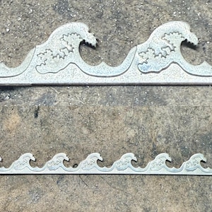 以下が含まれることがあります： 2つの金属製の波型の装飾品。上部のパーツには2つの完全な波があり、下部のパーツには6つの小さな波があります。波は、詳細でテクスチャ加工されたデザインのライトブルーです。背景はテクスチャ加工されたグレーの表面です。