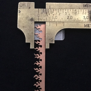 May include: A close-up of a metal ruler measuring a copper-colored metal strip with a flame design. The ruler is marked in inches and the measurement is 1.75 inches.