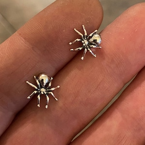 Puede incluir: Dos peque&ntilde;os pendientes de ara&ntilde;a de color plateado. Cada pendiente presenta un cuerpo redondeado y ocho delicadas patas. Los pendientes se muestran sobre un fondo neutro.