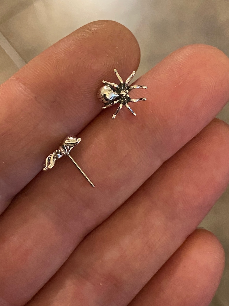 Puede incluir: Un par de pendientes de plata. Un pendiente es una ara&ntilde;a con ocho patas y un cuerpo redondeado. El otro pendiente es un peque&ntilde;o dise&ntilde;o estilizado. Los pendientes se muestran sobre una superficie marr&oacute;n claro.