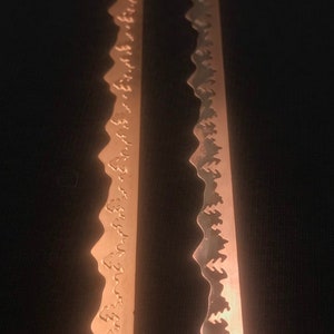 May include: Two close-up images of a metal saw blade with a decorative pattern of small trees along the edge. The saw blade is a light copper colour.