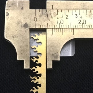 May include: A close-up of a metal ruler measuring a gold-coloured metal strip with a flame-like pattern. The ruler shows measurements in inches and centimetres.