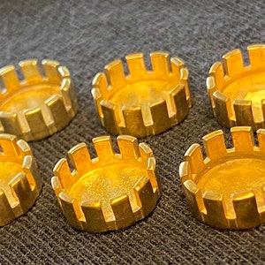 May include: Six small, gold-colored metal components with a circular design. Each component has a flat base and a notched, crown-like edge. The items are arranged on a dark, textured surface.