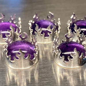 May include: Five silver crown rings with a purple, oval stone. The crowns have a flame design around the edges. The rings are displayed on a metallic surface. The rings are suitable for jewellery.