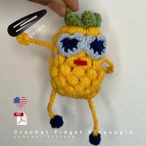 May include: A yellow crocheted pineapple figure with green leaves, light blue and dark blue eyes, a red mouth, and pink cheeks. It has thin yellow arms and legs with dark blue feet. A black hair clip is attached to the top, labeled Click-clack.
