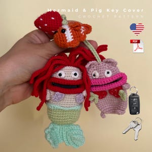 May include: A crochet pattern for a mermaid and pig key cover featuring text that reads Mermaid & Pig Key Cover Crochet Pattern. The design includes a red-haired mermaid, a pink pig, and a small orange fish with a red strawberry attached.