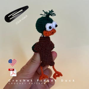 May include: A brown and dark green crochet duck toy with white eyes and orange feet. The duck features a tuft of green yarn on its head and an orange beak. A black hair clip is shown in the corner with the text Click-clack and Crochet Fidget Duck.