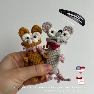 Może przedstawiać: Dwie szydełkowe zabawki typu fidget: kot i mysz. Kot jest w kolorze musztardowym z różowym szalikiem, a mysz jest szara z czerwonym szalikiem. Na zdjęciu znajduje się również czarna spinka do włosów z napisem "Click-clack". Tekst "Crochet Cat & Mouse Fidget Toy Pattern" znajduje się na dole.