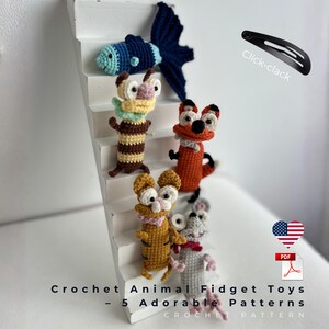 Può includere: Una collezione di cinque giocattoli fidget a forma di animale all'uncinetto, tra cui un pesce, una volpe e un topo, esposti su una scala bianca. I giocattoli sono in vari colori, con il testo "Crochet Animal Fidget Toys - 5 Adorable Patterns" in basso. È visibile anche un fermaglio nero.