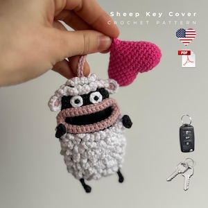 May include: A handmade crochet sheep key cover with a pink heart-shaped charm. The sheep features white wool, black details, and a pink mouth. The text "Sheep Key Cover Crochet Pattern" is visible. Includes a car key and two metal keys.