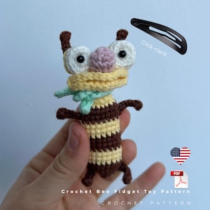 May include: A handmade crochet bee fidget toy with a brown and yellow striped body, white eyes, and a pink nose. The bee has a yellow scarf and a green scarf. The text "Crochet Bee Fidget Toy Pattern" is at the bottom. A black hair clip is in the image.