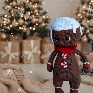 May include: A crocheted gingerbread man doll with a white icing head, red scarf, and decorative buttons. The brown doll has white accents on its arms and legs. The background includes Christmas trees and wrapped gifts, suggesting a festive holiday theme.