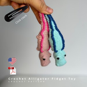 May include: Two crochet alligator fidget toys, one pink with a magenta spine and one light blue with a dark blue spine, each featuring black bead eyes. A black metal hair clip is included, labeled with the text Click-clack.