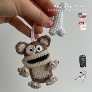 May include: Crocheted monkey key cover with a matching bone-shaped charm. The monkey is brown and beige with black accents. The image also shows a car key fob and two metal keys. Text on the image reads "Dog Key Cover Crochet Pattern".