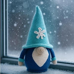 Può includere: Un gnomo all'uncinetto fatto a mano con un cappello a punta blu ornato da un fiocco di neve bianco. Lo gnomo ha un corpo blu, barba color crema e piccole braccia e piedi. Lo sfondo mostra una scena innevata, suggerendo un tema invernale.