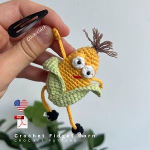 May include: A handmade crochet corn character with googly eyes, a red smile, and brown hair. The corn is yellow with green husks and black feet. A black hair clip is in the upper left corner. The text "Click-clack" is on the clip. The text "Crochet Fidget Corn Crochet Pattern" is at the bottom.