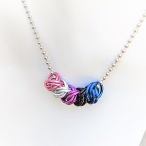 May include: A silver chain necklace with a multi-colored woven charm. The charm is made of pink, white, black, and blue metal rings.