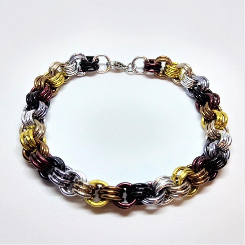 Op de afbeelding: Een veelkleurige metalen kettingarmband met afwisselende gouden, zilveren, zwarte en bordeauxrode schakels. De armband is gemaakt van een geweven kettingpatroon.