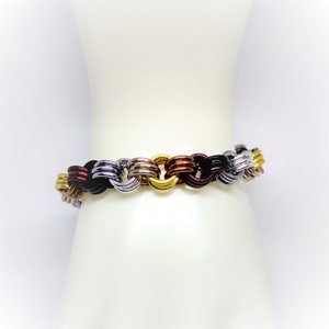 Op de afbeelding: Een veelkleurige armband gemaakt van met elkaar verbonden metalen ringen. De armband is gemaakt van zwarte, zilveren, gouden en koperkleurige ringen.