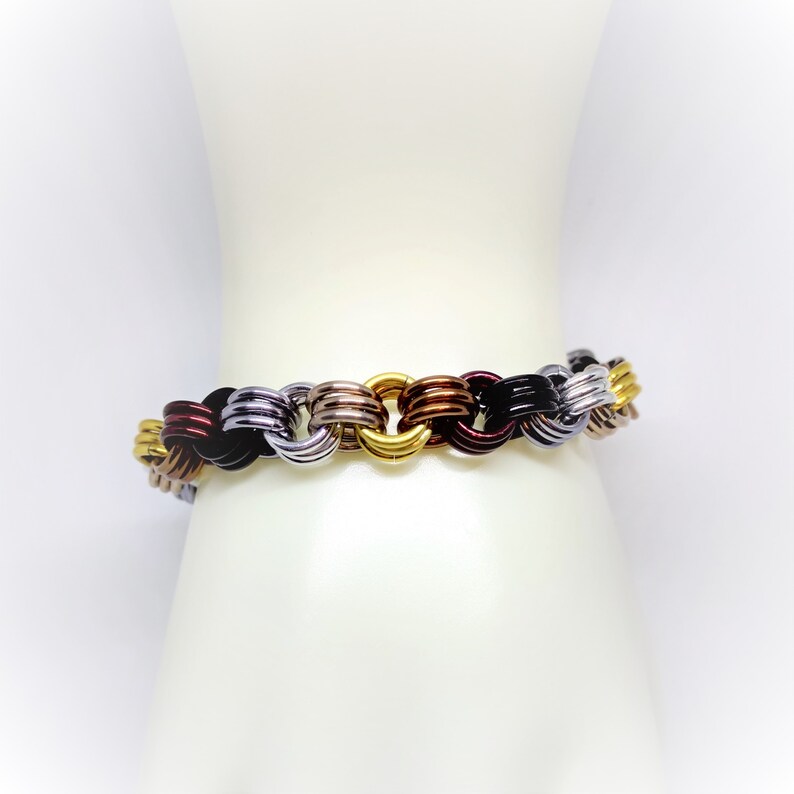 Op de afbeelding: Een veelkleurige armband gemaakt van met elkaar verbonden metalen ringen. De armband is gemaakt van zwarte, zilveren, gouden en koperkleurige ringen.