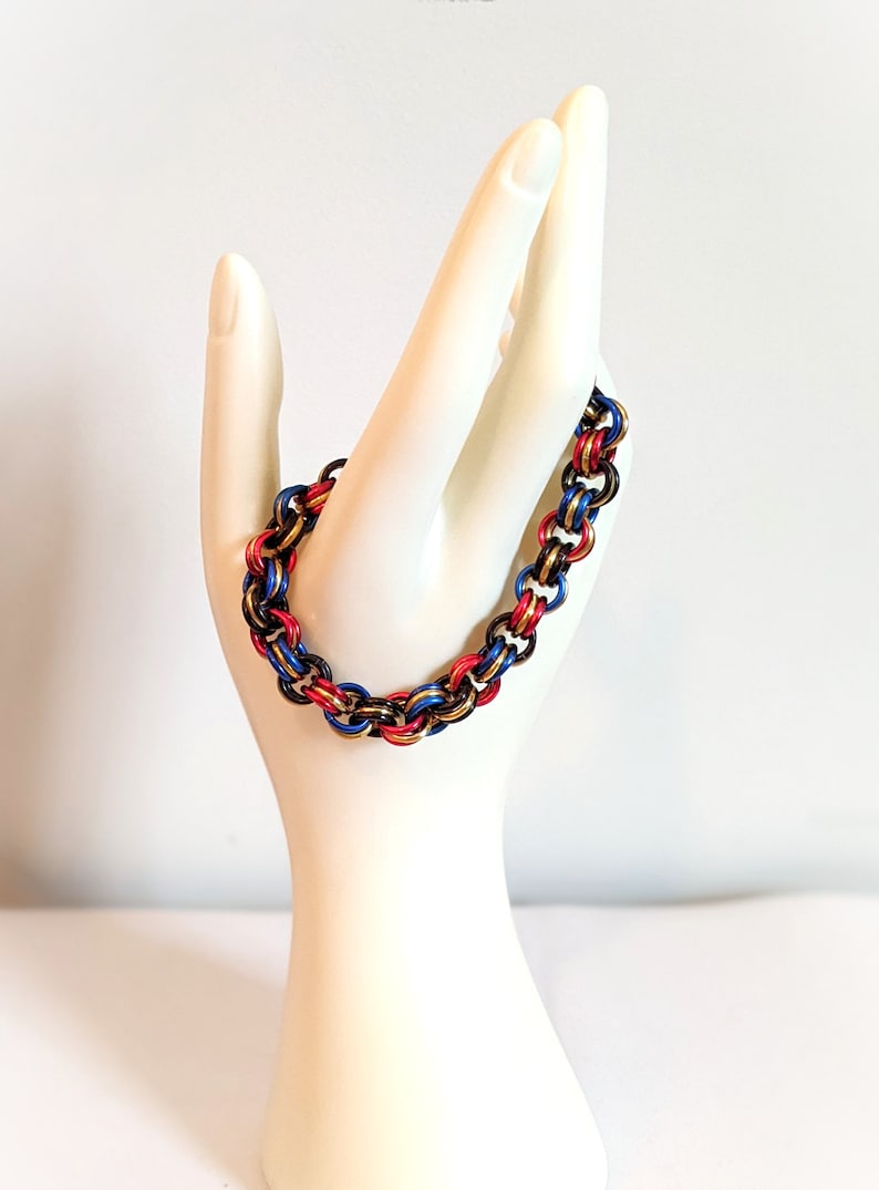 K&ouml;nnte beinhalten: Ein goldfarbenes, rotes und blaues Kettenhemdarmband. Das Armband besteht aus kleinen Metallringen, die miteinander verflochten sind, um ein Kettenhemdmuster zu erzeugen.
