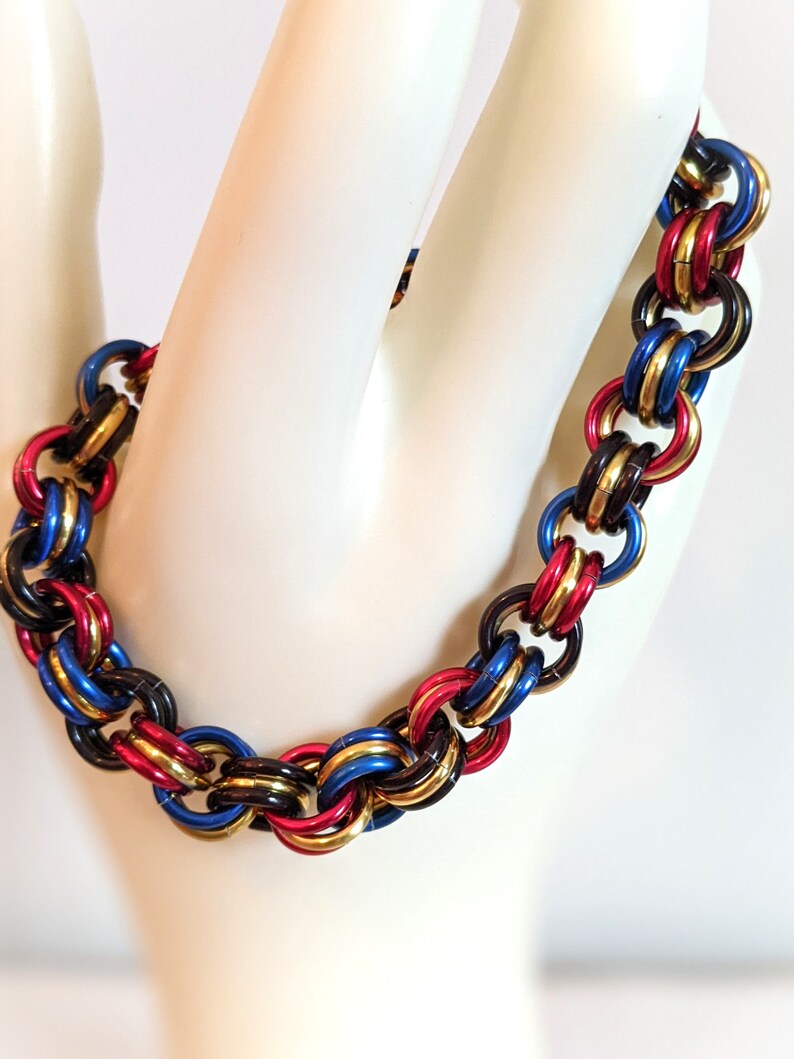 K&ouml;nnte beinhalten: Ein mehrfarbiges Armband aus Metallringen. Die Ringe sind schwarz, gold, rot und blau. Das Armband befindet sich an einer wei&szlig;en Mannequinhand.