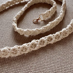 Puede incluir: Correa de macramé color crema con un cierre dorado. La correa está hecha de cordón de algodón tejido y presenta un patrón decorativo. La correa se muestra sobre una superficie texturizada de color neutro.