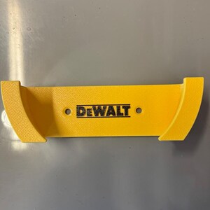 May include: A yellow plastic tool holder with the black "DEWALT" logo. The holder has a rectangular centre with curved ends and two mounting holes. The holder is designed to mount tools.