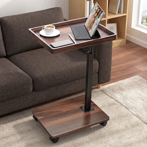May include: A dark wood-grain finish rolling laptop table with a black metal support post and base. The rectangular tabletop features a white ceramic coffee cup, a smartphone, a stylus, and a tablet computer with an attached keyboard.