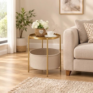 May include: A round side table with a gold metal frame, a flat top surface, and a lower beige fabric storage basket. The table holds a woven basket filled with white flowers and a white ceramic teacup on a matching saucer.