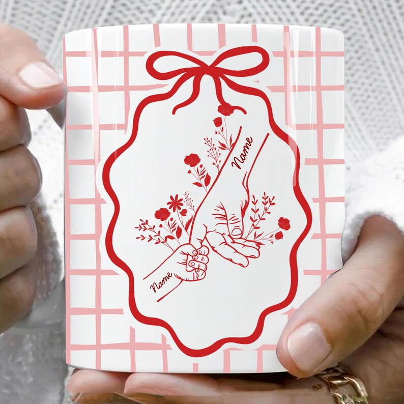 Op de afbeelding: Witte keramische mok met een rood bloemmotief met een ouderhand die de hand van een kind vasthoudt, met het woord "Name" in script. De mok heeft een roze rasterpatroon achtergrond en een rode strik aan de bovenkant.