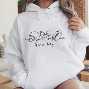 Pode incluir: Moletom branco com um desenho de linha preta de um capacete de futebol americano, um la&ccedil;o e uma bola de futebol. As palavras "Game Day" est&atilde;o escritas abaixo. O moletom tem capuz com cord&atilde;o e um bolso frontal.