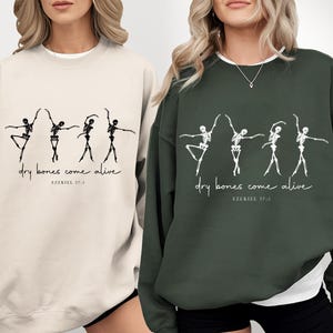 May include: Two sweatshirts, one cream and one dark green, each featuring a graphic of dancing skeletons. The text "dry bones come alive" and "Ezekiel 37:3" is printed below the skeletons. The sweatshirts are casual wear.