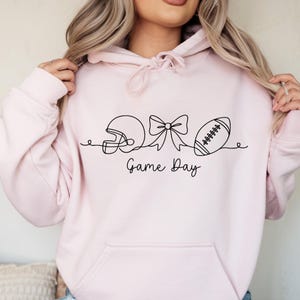 Pode incluir: Moletom rosa claro com um desenho de linha preta de um capacete de futebol americano, um la&ccedil;o e uma bola de futebol. As palavras "Game Day" est&atilde;o escritas abaixo. O moletom tem um bolso frontal e um capuz.