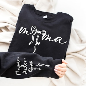 May include: Black sweatshirt with the word "mama" in white script, featuring a bow design. The sleeve displays the names "Megan Aiden" in white script, also with a bow design. The sweatshirt is folded on a cream-coloured fabric.