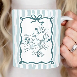 Op de afbeelding: Witte keramische mok met een lichtblauwe en witte gestreepte achtergrond. De mok heeft een teal-kleurig frame met een strik aan de bovenkant, met daarin een ontwerp van handen die elkaar vasthouden, met het woord "Name" op elke hand.