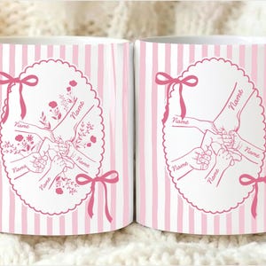 Op de afbeelding: Twee witte keramische mokken met roze verticale strepen. Elke mok heeft een roze ovaal ontwerp met handen die elkaar vasthouden, bloemaccenten en het woord "Name". Roze strikken sieren het ontwerp.
