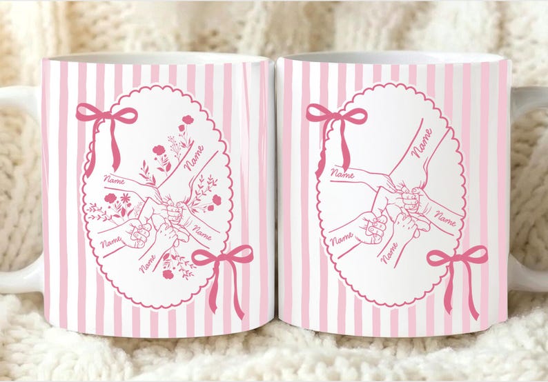 Op de afbeelding: Twee witte keramische mokken met roze verticale strepen. Elke mok heeft een roze ovaal ontwerp met handen die elkaar vasthouden, bloemaccenten en het woord "Name". Roze strikken sieren het ontwerp.