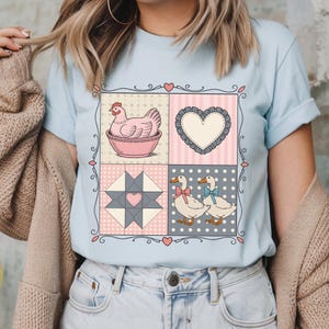 以下が含まれることがあります： パッチワークデザインのライトブルーTシャツ。ピンクのボウルに入った鶏、ハート、キルトブロック、リボンをつけた2羽のアヒルが、ハートの装飾的なボーダーの中に描かれています。