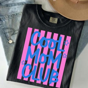 May include: Black t-shirt with the text "COOL MOM CLUB" in blue, set against pink vertical stripes. The shirt is paired with blue jeans and a tan hat.
