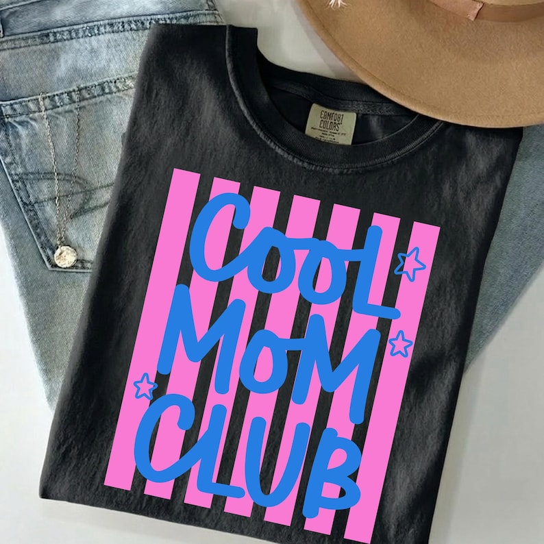 May include: Black t-shirt with the text "COOL MOM CLUB" in blue, set against pink vertical stripes. The shirt is paired with blue jeans and a tan hat.