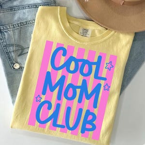 May include: A light yellow t-shirt with the words "Cool Mom Club" in blue script and pink vertical stripes. The shirt is folded on top of blue jeans and a brown hat.
