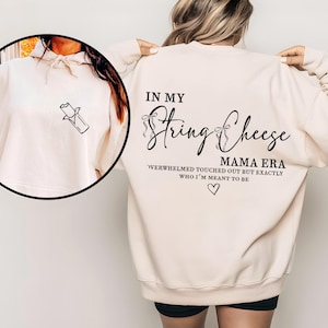 May include: A cream-coloured sweatshirt featuring the text "IN MY String Cheese MAMA ERA" in black script. A small graphic of a candy bar is on the front. The back of the sweatshirt also has text, including "WERWHELMED TOUCHED OUT BUT EXACTLY WHO I'M MEANT TO BE" and a heart symbol.