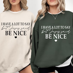 May include: Two crewneck sweatshirts, one cream with black text and one dark green with white text. Both feature the phrase "I HAVE A LOT TO SAY but Jesus said BE NICE" in a mix of block and script fonts, finished with a small heart icon.