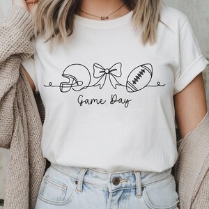 Pode incluir: Camiseta creme com um desenho a tra&ccedil;o preto de um capacete de futebol americano, um la&ccedil;o e uma bola de futebol. As palavras "Game Day" est&atilde;o escritas abaixo. A camisa &eacute; usada com jeans azuis.